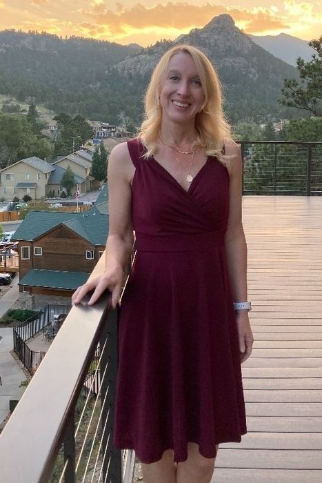 A reviewer stands on a balcony in a stunning v-neck burgundy dress, with a breathtaking mountain landscape serving as the perfect backdrop.
