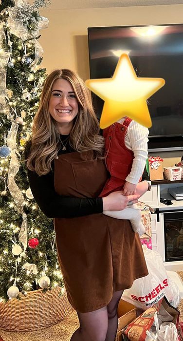 A reviewer smiles while wearing a brown corduroy jumper, holding a small child in front of a festive Christmas tree, surrounded by holiday gift bags and boxes in a cozy living room