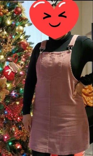 A person in a pink corduroy pinafore dress and a black turtleneck stands beside a beautifully decorated Christmas tree