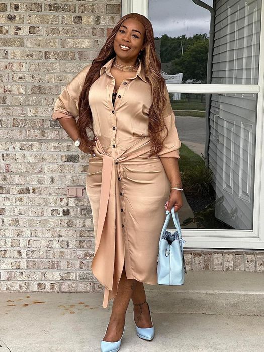A person models a fashionable khaki button-up satin dress paired with blue heels and a matching handbag, standing outside a charming brick building.
