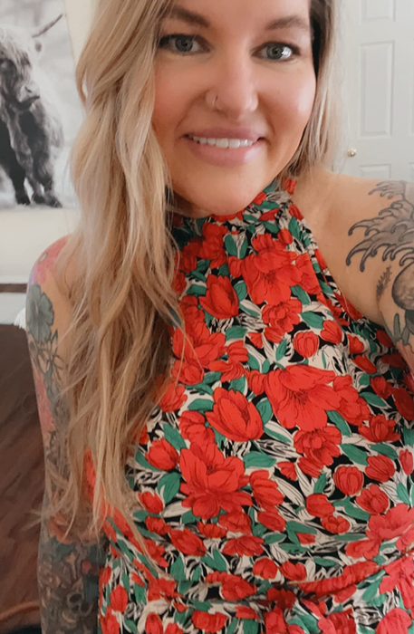 Woman in a floral dress poses for a selfie, tattooed arms visible, with wall art in the background