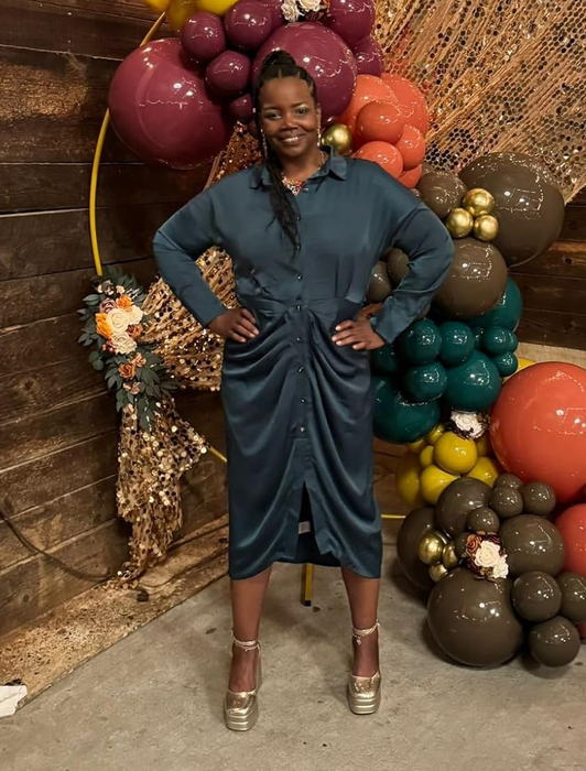 A person stands smiling in a chic dark green ruched dress, positioned next to a decorative balloon arrangement, with wooden walls and warm ambient lighting creating a cozy atmosphere.