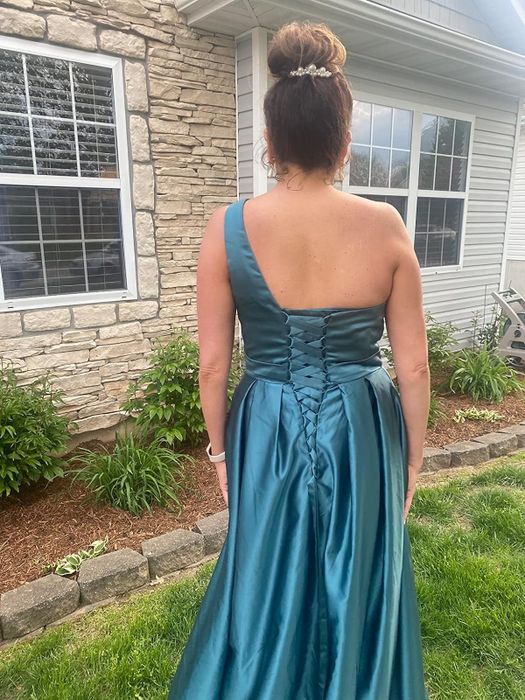 A reviewer stands in a backyard garden wearing a blue one-shoulder satin gown, styled with an elegant updo. The image highlights the dress’s corseted back design