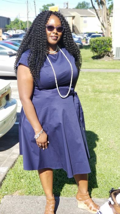 A person outdoors, wearing a chic knee-length dress, sunglasses, and a long pearl necklace, poses beside parked cars