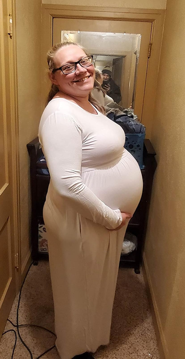 A pregnant reviewer smiles in a hallway, wearing a flowing white maxi dress