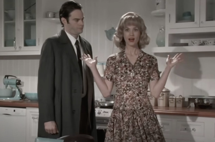 A woman in a vintage floral dress gestures dramatically in a retro kitchen, while a man in a suit observes