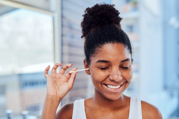 A person with their eyes closed, smiling softly, while gently using a cotton swab on their face in a bathroom setting