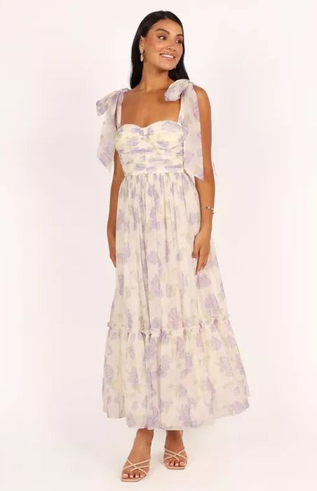 Model wearing a floral maxi dress with bow straps and a ruffled hem, accessorized with hoop earrings and sandals, smiling toward the side.