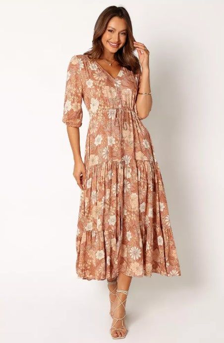 A model showcasing a floral-patterned, long, flowing dress with three-quarter sleeves, posing and smiling. The model completes the look with strappy sandals and a bracelet.