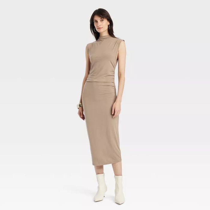 A model in a sleeveless, form-fitting ankle-length dress, paired with ankle boots, poses against a plain backdrop.