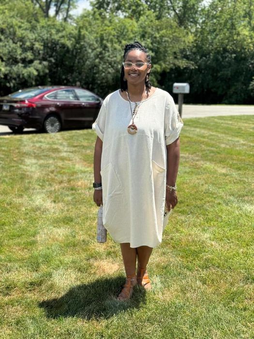 A reviewer stands on grass in a relaxed, light-colored dress paired with sandals and accessories. She holds a small clutch and smiles at the camera