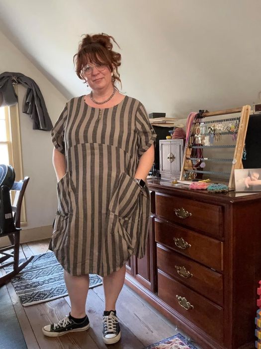 reviewer in a loose, striped dress with pockets, wearing black sneakers, standing in a room with a wooden dresser and various jewelry pieces