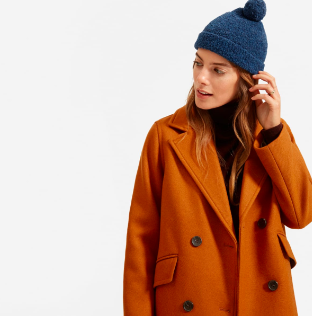 A model is seen wearing a dark blue teddy beanie with a burnt orange trench coat