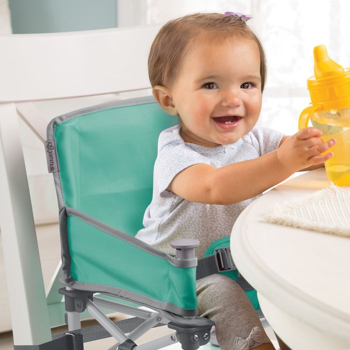 A toddler seated in a booster seat attached to a dining chair, holding a bottle
