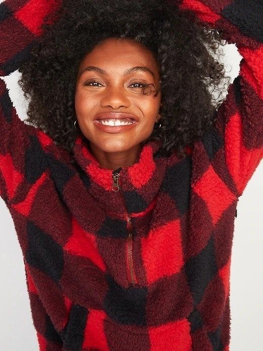 A woman playfully raising her arms in a half-zip red and black plaid sweatshirt.