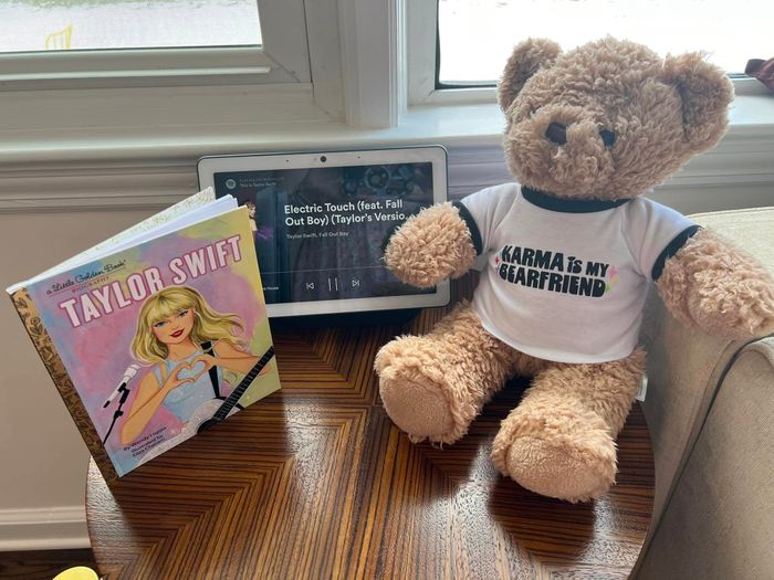 A Taylor Swift Little Golden Book, a tablet playing “Electric Touch”, and a teddy bear wearing a “KARMA IS MY BOYFRIEND” shirt