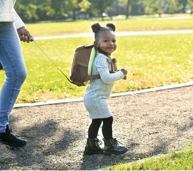 A smiling toddler in a sweater dress and boots, wearing a backpack, is held by an adult’s hand while outdoors.