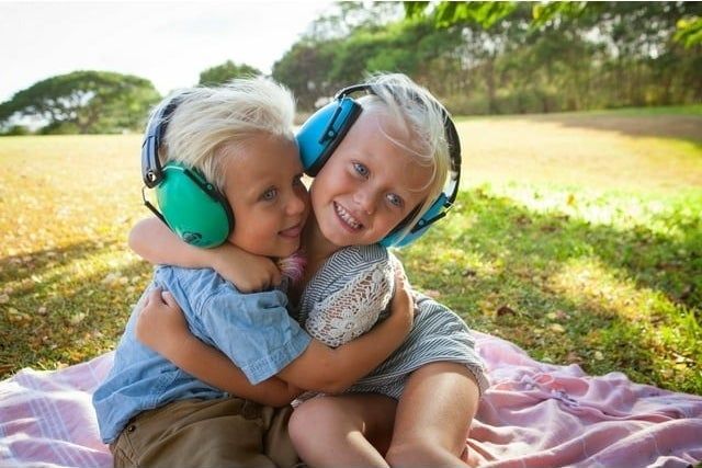 Two kids wearing noise-canceling headphones share a cozy moment on a picnic blanket outside.