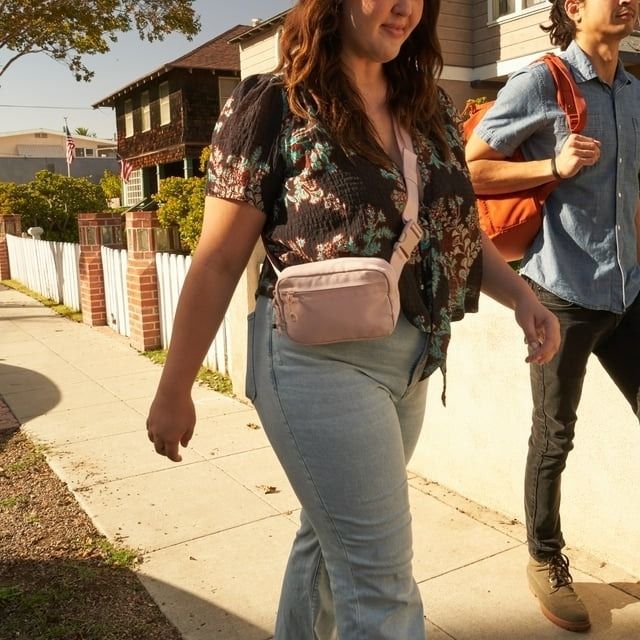 A person in a floral top and jeans wearing a pink waist bag walks on a sidewalk, with another person visible in the background.