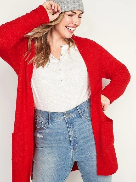 A woman in a long open-front red sweater, paired with a white t-shirt tucked into blue jeans, topped off with a grey beanie.