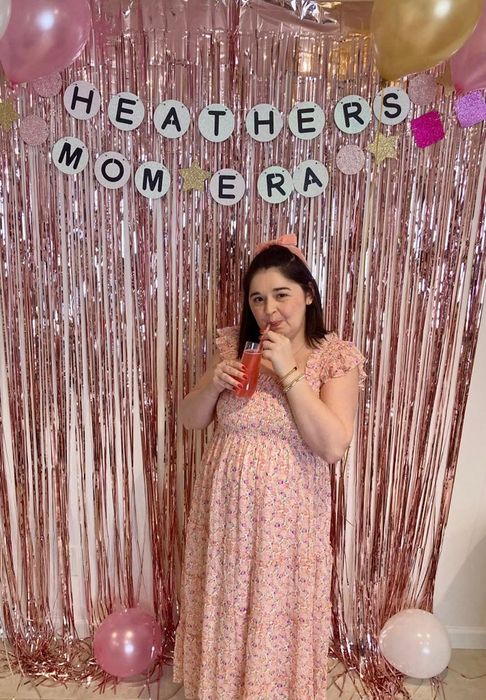 A woman in a floral dress posing in front of a pink foil backdrop with a banner reading "HEATHERS MOM ERA", holding a drink