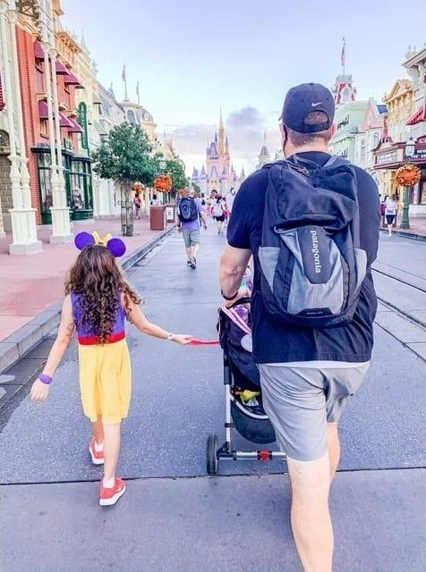 A man and child stroll down a theme park’s main street; the child wears mouse ears, and the man carries a backpack.