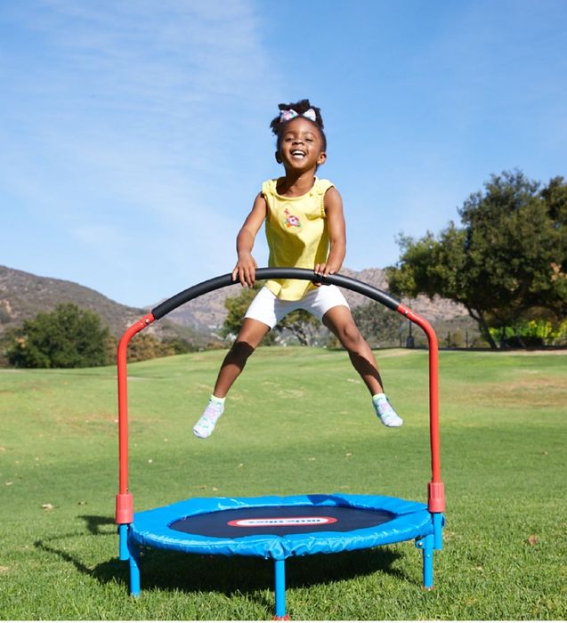 A child bounces joyfully on an outdoor trampoline, holding the handlebar, in a sunny grassy area
