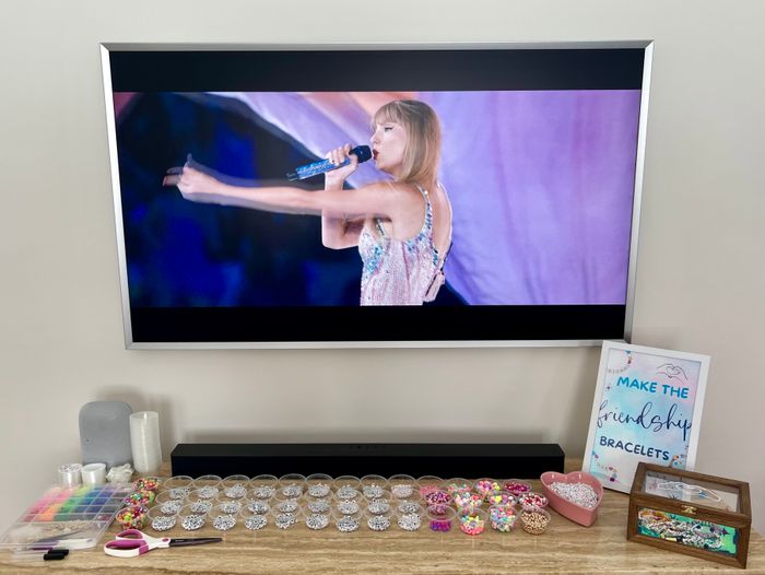 Taylor Swift performing on TV with a microphone, surrounded by jewelry-making supplies on a table in the foreground