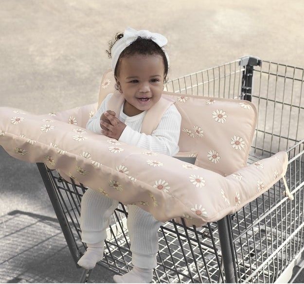 A smiling toddler sits comfortably in a shopping cart, supported by a floral-patterned cushion insert