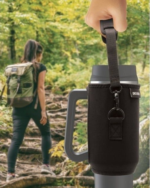 A person hiking in the background, with another person’s hand holding a travel mug in the foreground.