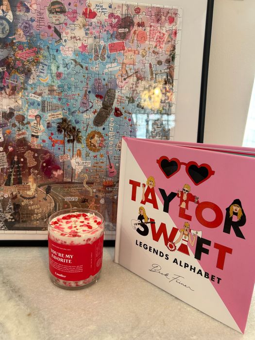 A candle with text, the Taylor Swift 'Legends Alphabet' book, and a completed jigsaw puzzle displayed on a counter
