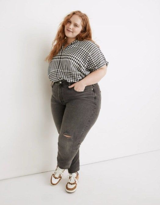 plus-sized model wearing vintage-wash gray jeans with a small tear at the knee, paired with brown and white sneakers and a black-and-white gingham shirt
