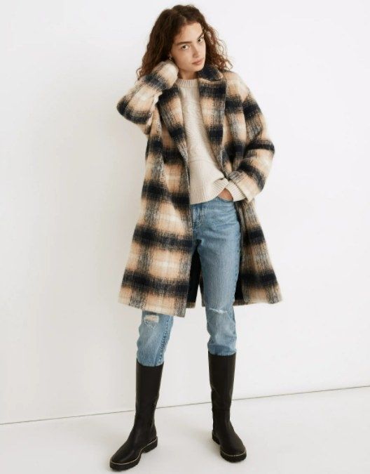 A model styled in a plaid black and yellowish coat, paired with jeans, black boots, and a cozy cream-colored sweater.
