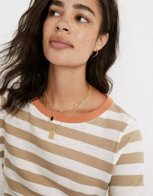 model styling the necklace set with a short chain, a medium-length black stone necklace, and a long brass pendant necklace layered over a brown and white striped shirt