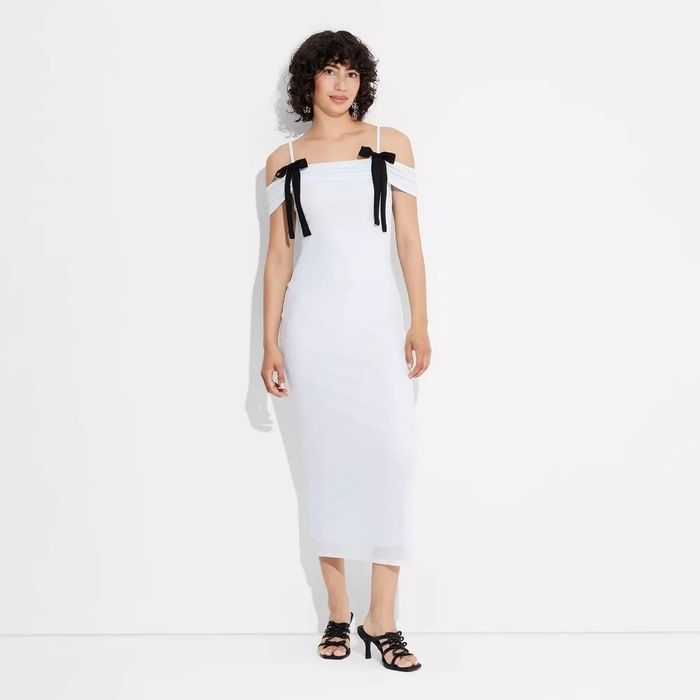 The model is dressed in an off-shoulder white dress, featuring black ribbon details and paired with black strappy heels.
