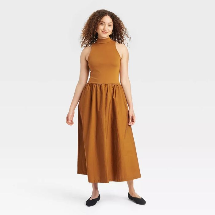The model is wearing a sleeveless, high-neck, ankle-length dress paired with black flats.