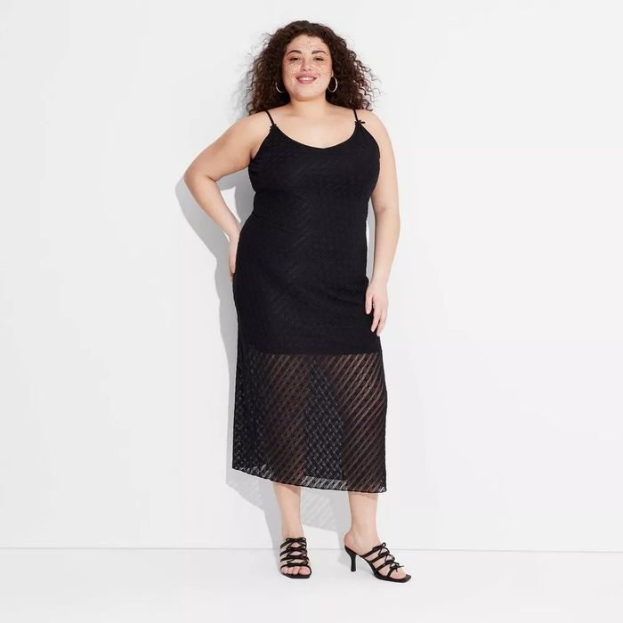 The model is wearing a sleeveless, midi-length black dress with sheer detailing at the bottom, paired with strappy black high heels.