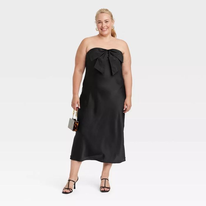 The model is wearing a black strapless dress with a large bow detail, holding a small clutch and black heels.