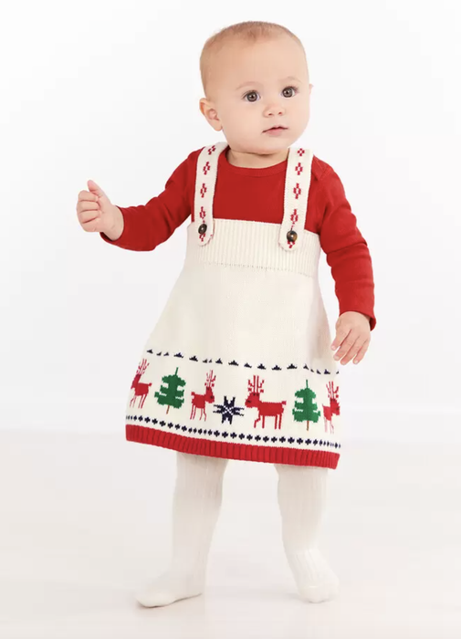 A baby in a white sweater jumper dress featuring holiday trees and deer designs.