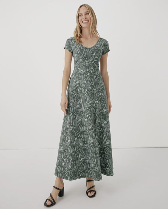 A woman in a green patterned maxi dress with short sleeves and black sandals, smiling in a studio setting for shopping content