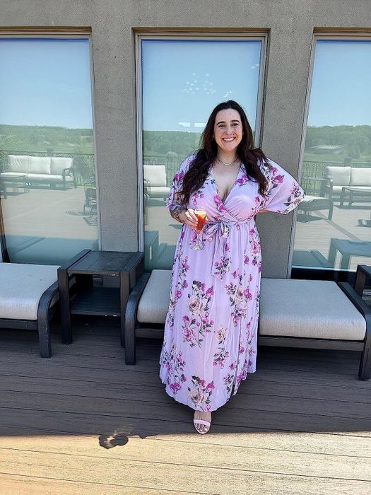 A reviewer wearing a three-quarter sleeve wrap-front purple dress adorned with pink flowers, highlighting its charm and versatility.