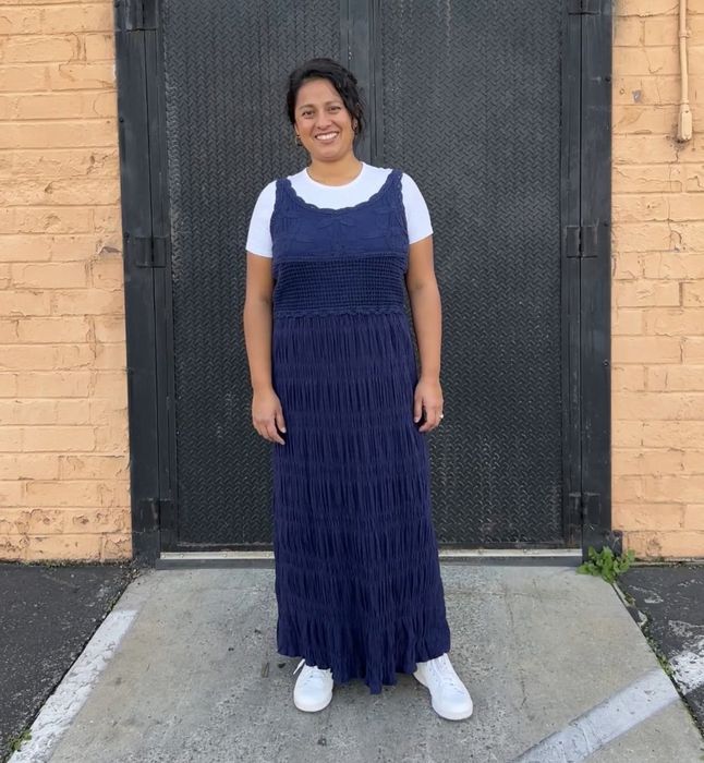 A woman wearing a tiered blue dress and white sneakers smiles brightly in front of a doorway.