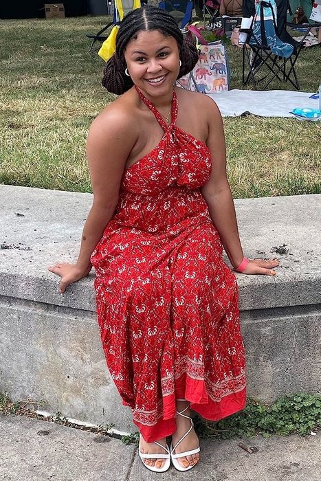 Another reviewer in a red patterned sundress and white sandals, smiling while seated on a ledge at an outdoor event