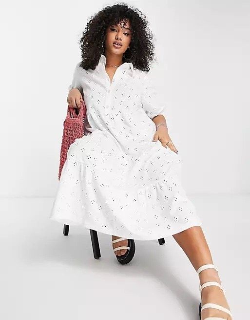 A model dressed in a white dress and matching white shoes, sitting gracefully in a chair, accessorized with a pink handbag.