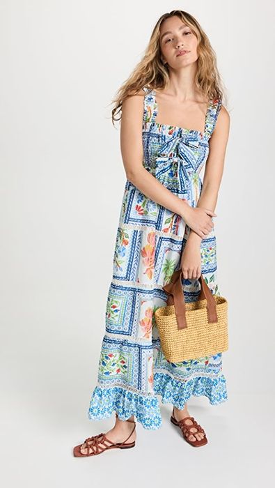 A model showcases a sleeveless maxi dress in a blend of blue, white, green, and orange hues, complete with a smocked bodice and playful bows.