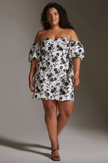 A model dons a white dress adorned with black floral appliques, showcasing a chic and elegant design.