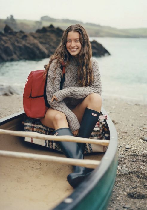 A person sitting in a canoe near a beach, dressed in a cable-knit sweater, rubber boots, and a backpack, smiling with a relaxed demeanor