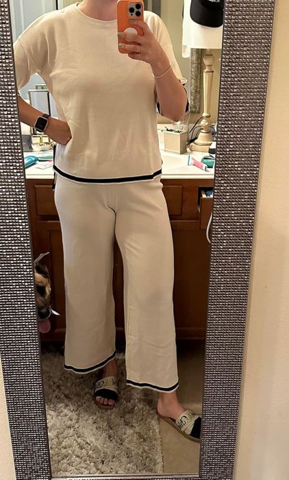 A reviewer wearing a casual top and pants poses in front of a mirror, with a dog partially visible in the background. The person is holding a phone