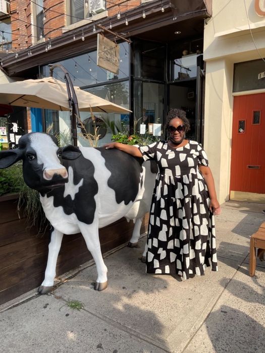 A reviewer posing next to a cow statue, wearing a black dress with white geometric patterns in a bright, urban setting