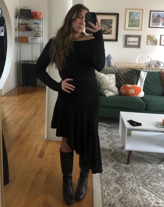 A reviewer takes a mirror selfie in a black dress and boots, standing in a living room adorned with framed pictures, a green couch, and a white rug